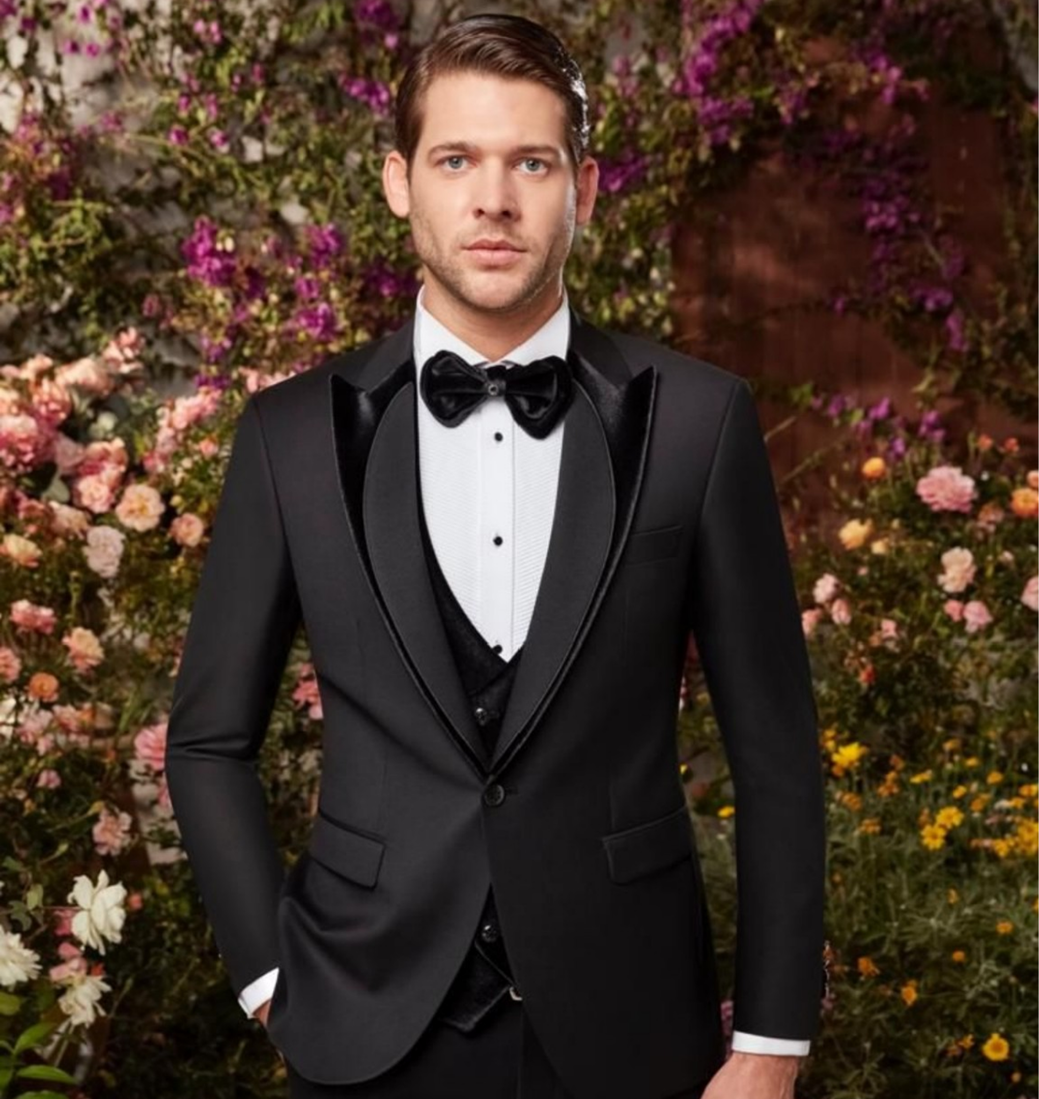 Man in tuxedo standing among flowers.